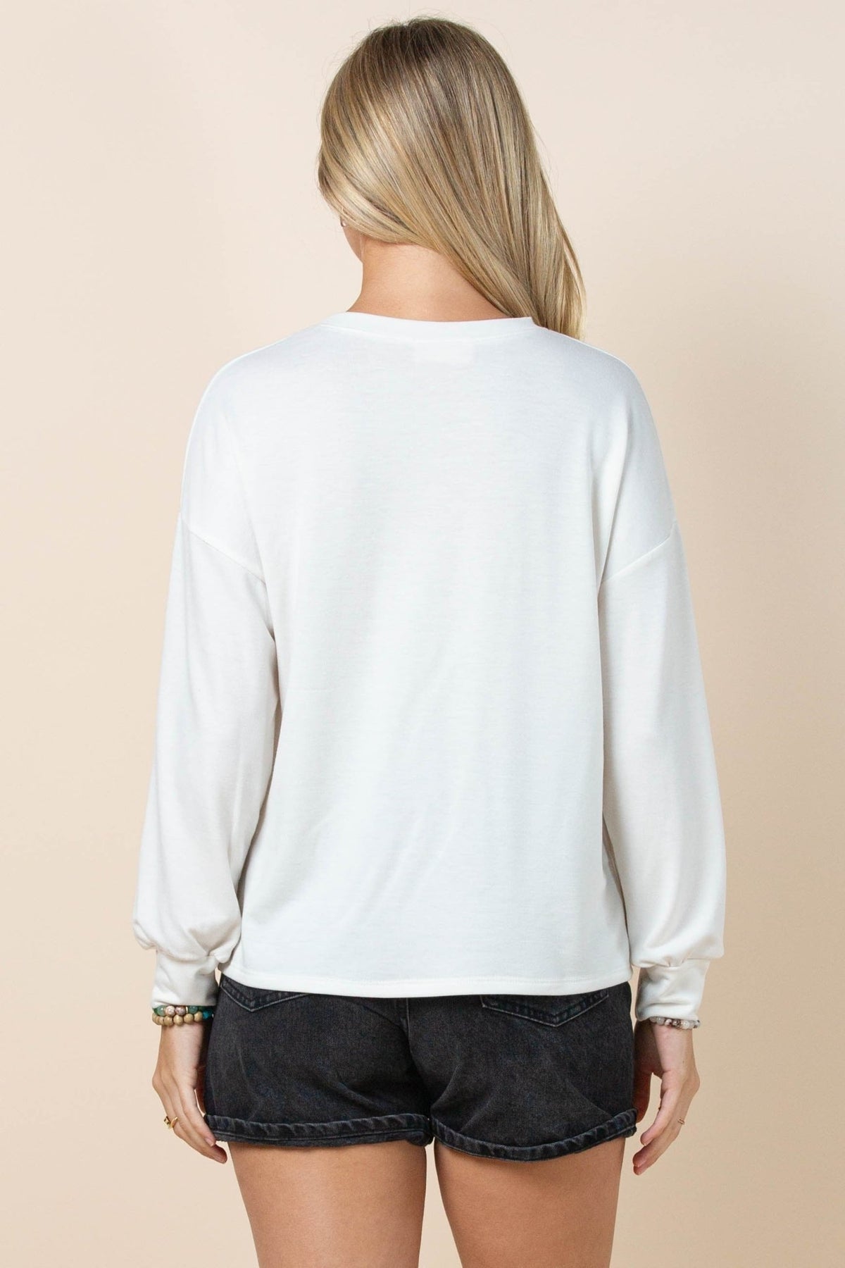 A woman shown from behind wearing a white long-sleeve crewneck sweatshirt and black denim shorts against a solid beige background.