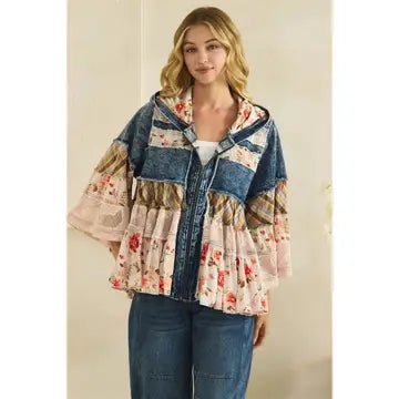 A woman models a bohemian-style patchwork hoodie jacket featuring a blue denim hood and sleeves with tiered ruffles and floral prints in pink, red, and beige, paired with blue jeans against a neutral beige wall.