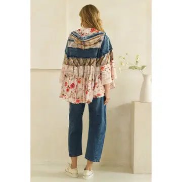 A woman shown from the back wearing a tiered bohemian-style patchwork blouse with blue denim and floral prints, paired with blue jeans and white sneakers, standing in a minimalist beige room.