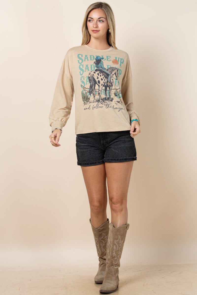 A woman modeling a beige long-sleeve graphic sweatshirt with a western-themed print, paired with dark denim shorts and brown cowboy boots, against a solid beige studio background.