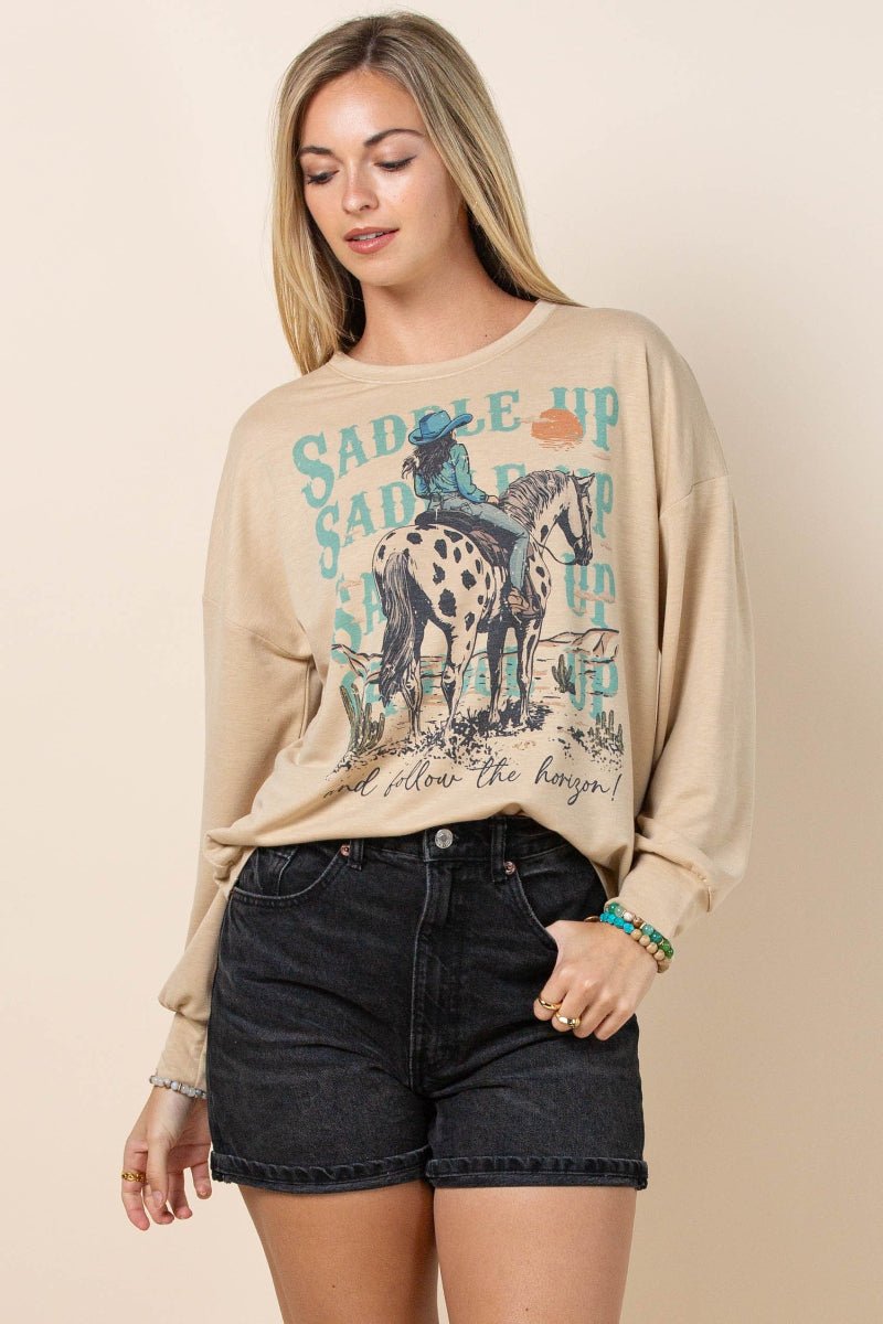 A woman modeling a beige crewneck sweatshirt with a western-themed graphic print of a cowboy on a dalmatian horse, paired with dark denim shorts against a solid beige background.