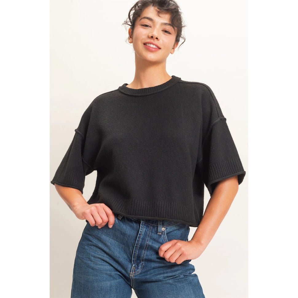 A woman wearing a black short-sleeve knit sweater and blue jeans, standing against a plain light background.