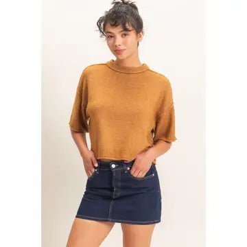 A woman modeling a mustard yellow short-sleeve knit top with a mock neck, paired with a dark blue denim mini skirt, against a plain off-white background.