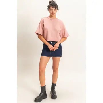 A woman modeling a light pink cropped t-shirt with short wide sleeves, paired with a dark denim mini skirt and black ankle boots, against a plain light beige studio background.