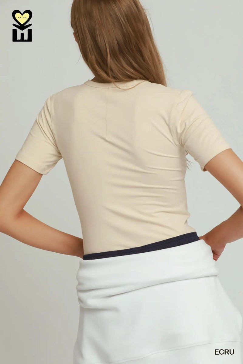 A woman shown from the back wearing a fitted short-sleeve beige top and a white skirt with a dark navy waistband, against a light gray studio background.