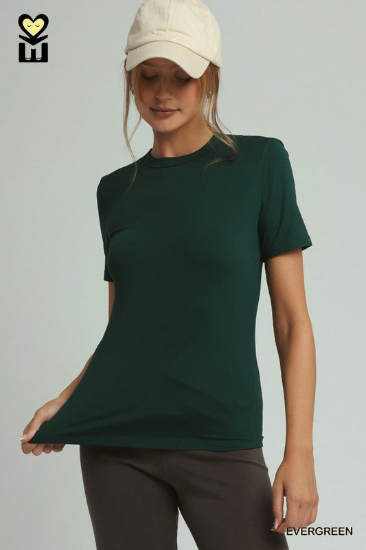 A woman wearing a dark forest green short-sleeve athletic t-shirt and dark gray leggings, holding the hem of the shirt to show its drape. She is wearing a cream-colored baseball cap.