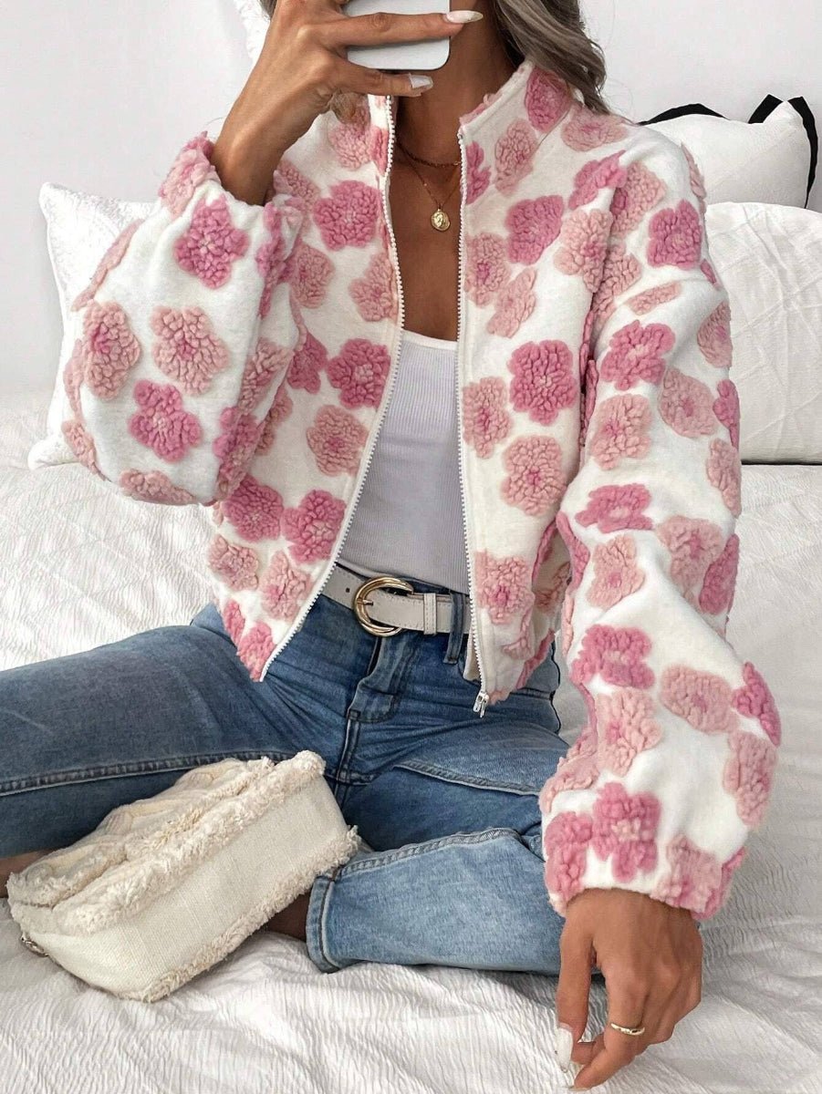 A woman sitting on a white bed wearing a white fuzzy bomber jacket with pink floral patterns, blue jeans, and a white tank top.
