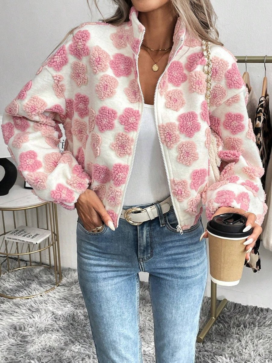 A woman wearing a fuzzy white zip-up jacket with pink pom-pom floral patterns, styled with a white tank top and blue jeans while holding a coffee cup.