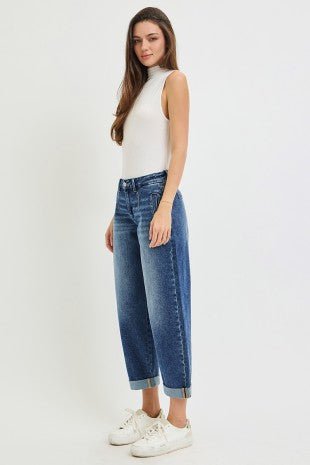 A woman modeling a pair of high-waisted, wide-leg cropped blue jeans with rolled cuffs, paired with a white sleeveless top and white sneakers, against a plain white studio background.