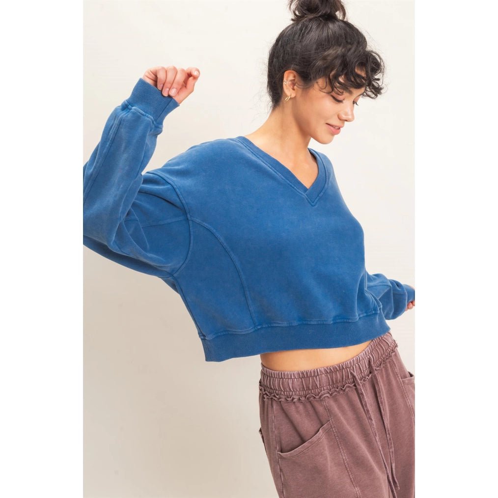 A woman wearing a cropped, long-sleeve V-neck sweatshirt in a washed medium blue color, paired with brown pants, posing against a plain off-white background.