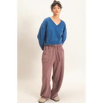 A model stands against a plain light beige studio background wearing a blue cropped V-neck sweatshirt and dusty rose wide-leg corduroy pants.