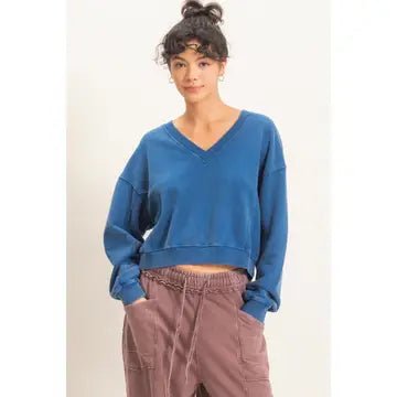 A woman wearing a cropped blue V-neck sweatshirt and dusty rose drawstring pants, standing against a plain off-white background.