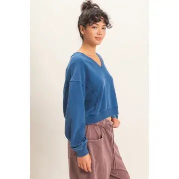 A woman wearing a blue cropped V-neck sweatshirt and dusty rose drawstring pants, photographed against a plain light beige studio background.