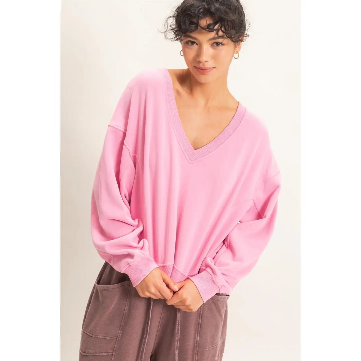 A woman wearing a pink oversized V-neck long-sleeve sweatshirt and brown corduroy pants, standing against a plain off-white background.