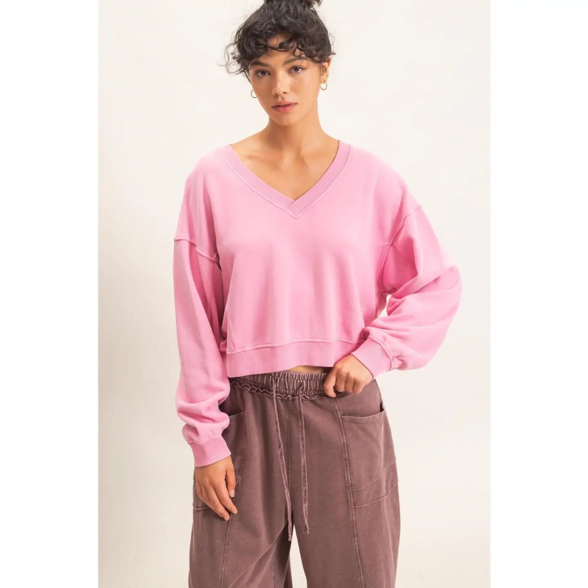 A woman modeling a light pink cropped V-neck sweatshirt with balloon sleeves, paired with brown drawstring pants, against a plain off-white studio background.
