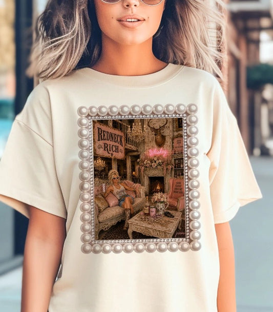 A woman wearing a cream-colored oversized t-shirt with a graphic print of a vintage scene framed by a pearl border, set against a blurred outdoor background.