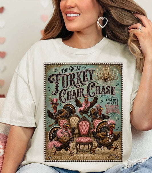 A woman wearing a cream-colored graphic t-shirt featuring a vintage-style illustration of turkeys chasing a pink chair with the text 'The Great Turkey and Chair Chase'.