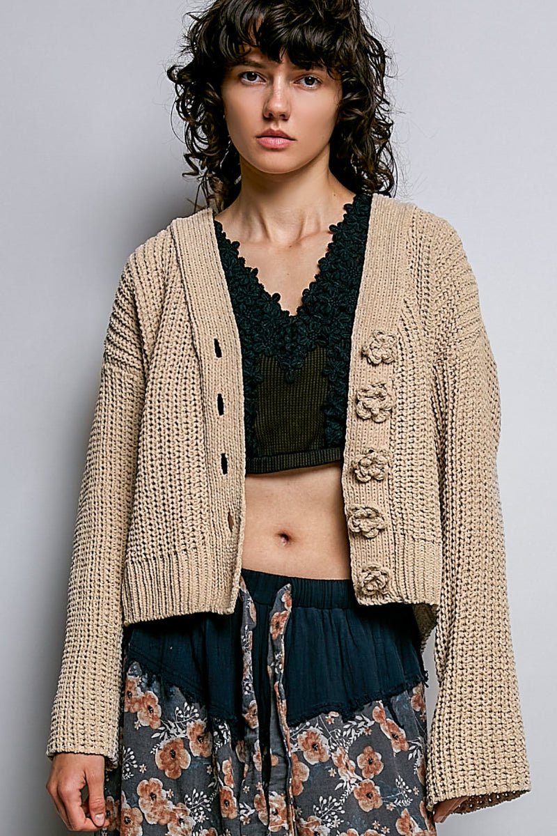 A model wearing a beige chunky knit cardigan with large decorative buttons, layered over a black lace-trim crop top and floral skirt, against a plain gray background.