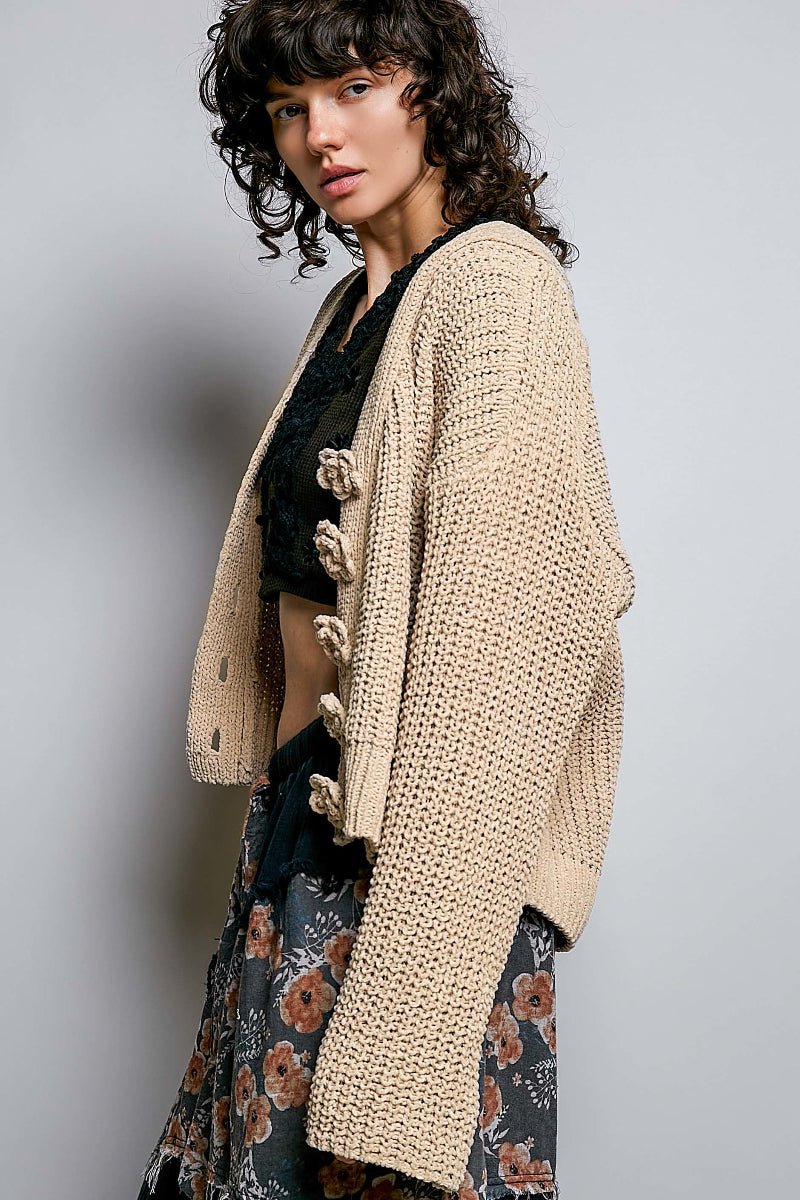 A woman wearing a chunky cream-colored open-front cardigan with large decorative buttons, styled over a black lace top and a dark floral skirt, set against a plain gray studio background.