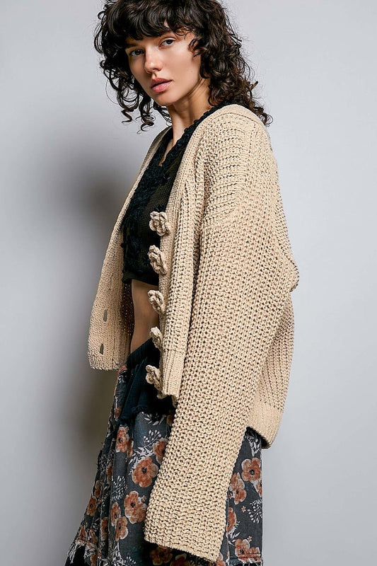 A woman wearing a chunky cream-colored open-front cardigan with large decorative buttons, styled over a black lace top and a dark floral skirt, set against a plain gray studio background.