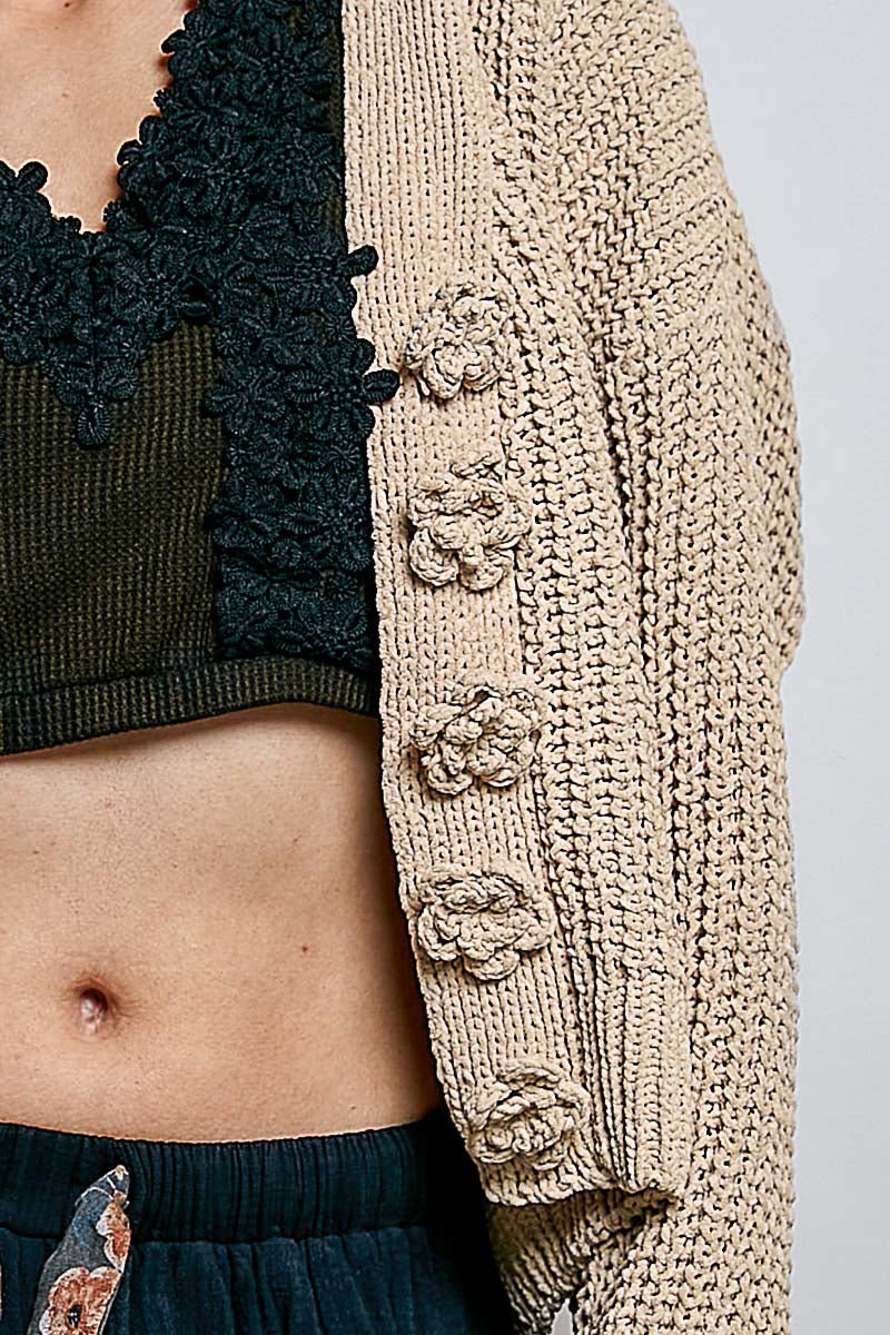 Close-up of a person wearing a chunky beige knit cardigan with large textured flower appliqués down the front, layered over a black lace-trim crop top and dark bottoms.