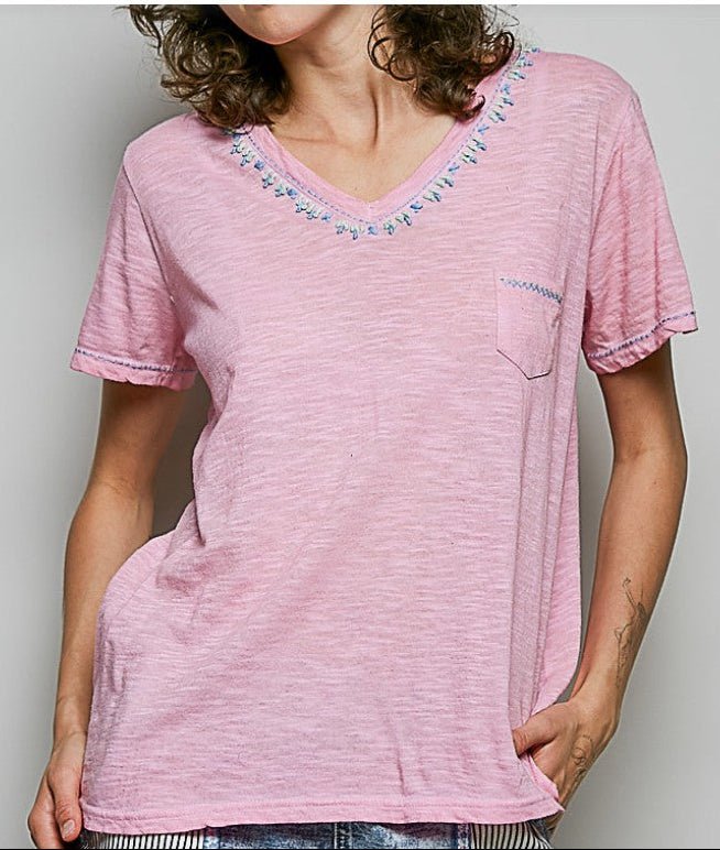 A woman wearing a light pink short-sleeve V-neck t-shirt with blue and white embroidered details around the neckline and a matching embroidered pocket.