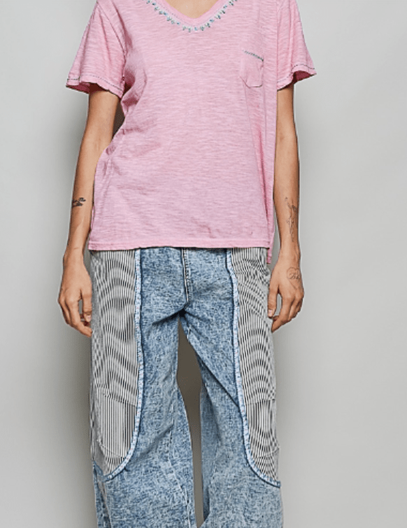 A model wearing a pink short-sleeve V-neck t-shirt with blue embroidered detailing along the neckline and a left chest pocket, paired with light blue denim jeans featuring striped patchwork panels on the legs, against a plain light gray background.