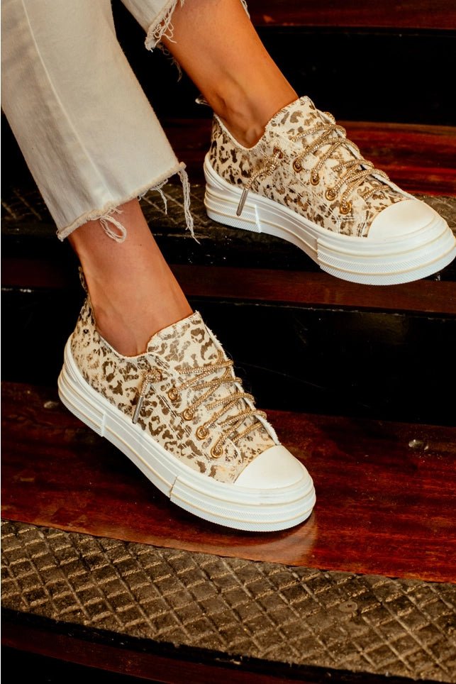 A person wearing beige and brown leopard-print low-top sneakers with chunky white platform soles, standing on dark wooden stairs.