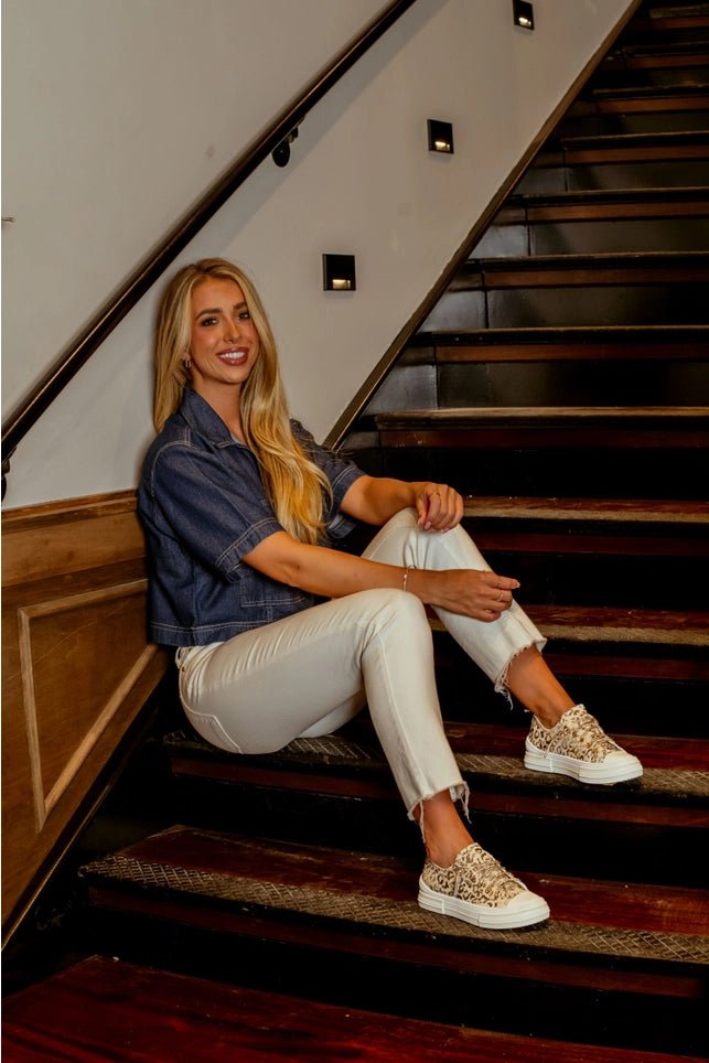 Woman sitting on a staircase wearing a denim jacket, white pants, and glittery sneakers.