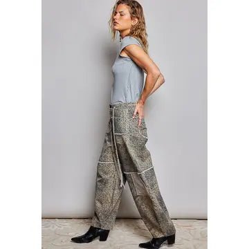 A woman modeling a pair of wide-leg, high-waisted pants with a snakeskin pattern in brown, beige, and gray tones, paired with a light blue sleeveless top and black boots, set against a plain gray studio background.