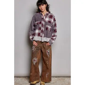A model wearing a vintage-style plaid button-up shirt jacket in gray and burgundy with a ruffled hem, paired with brown wide-leg pants featuring embroidered hearts and hot air balloon motifs, standing against a plain gray wall.