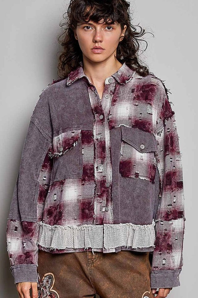 A model wearing a distressed, oversized button-up shirt jacket in a washed gray and deep burgundy plaid pattern, paired with brown pants, against a plain gray studio background.