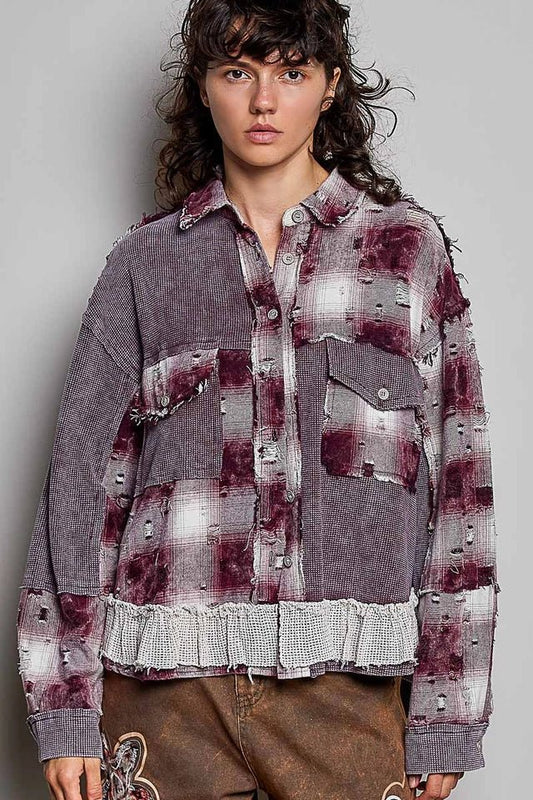 A model wearing a distressed, oversized button-up shirt jacket in a washed gray and deep burgundy plaid pattern, paired with brown pants, against a plain gray studio background.