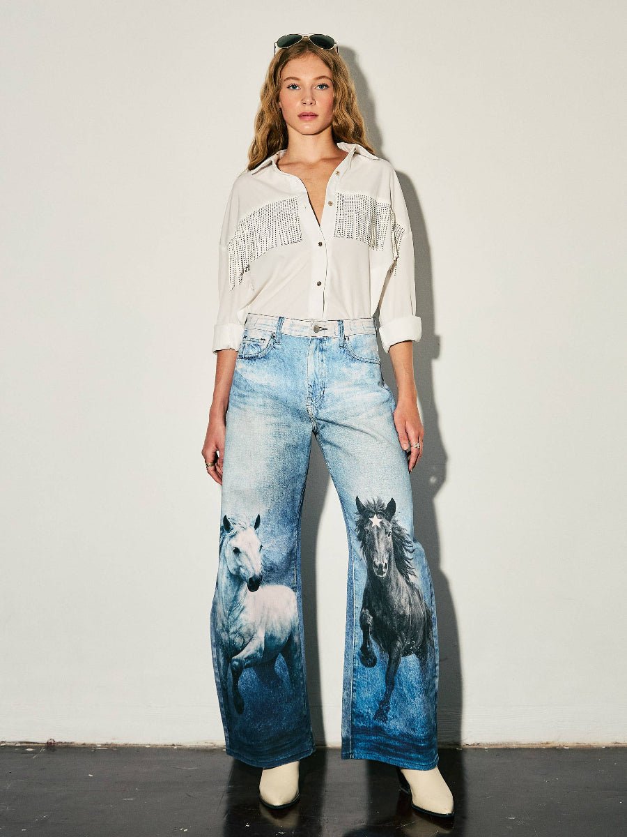 A model stands against a plain off-white wall wearing a white fringed button-up shirt and wide-leg blue jeans featuring printed images of two horses on the front legs, paired with white boots.