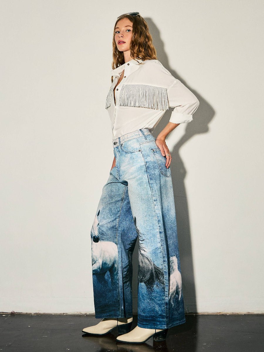 A model stands against a plain off-white wall wearing a white long-sleeve blouse with a silver fringe detail across the chest and wide-leg blue jeans featuring a printed white horse design. She is wearing off-white pointed-toe heels.