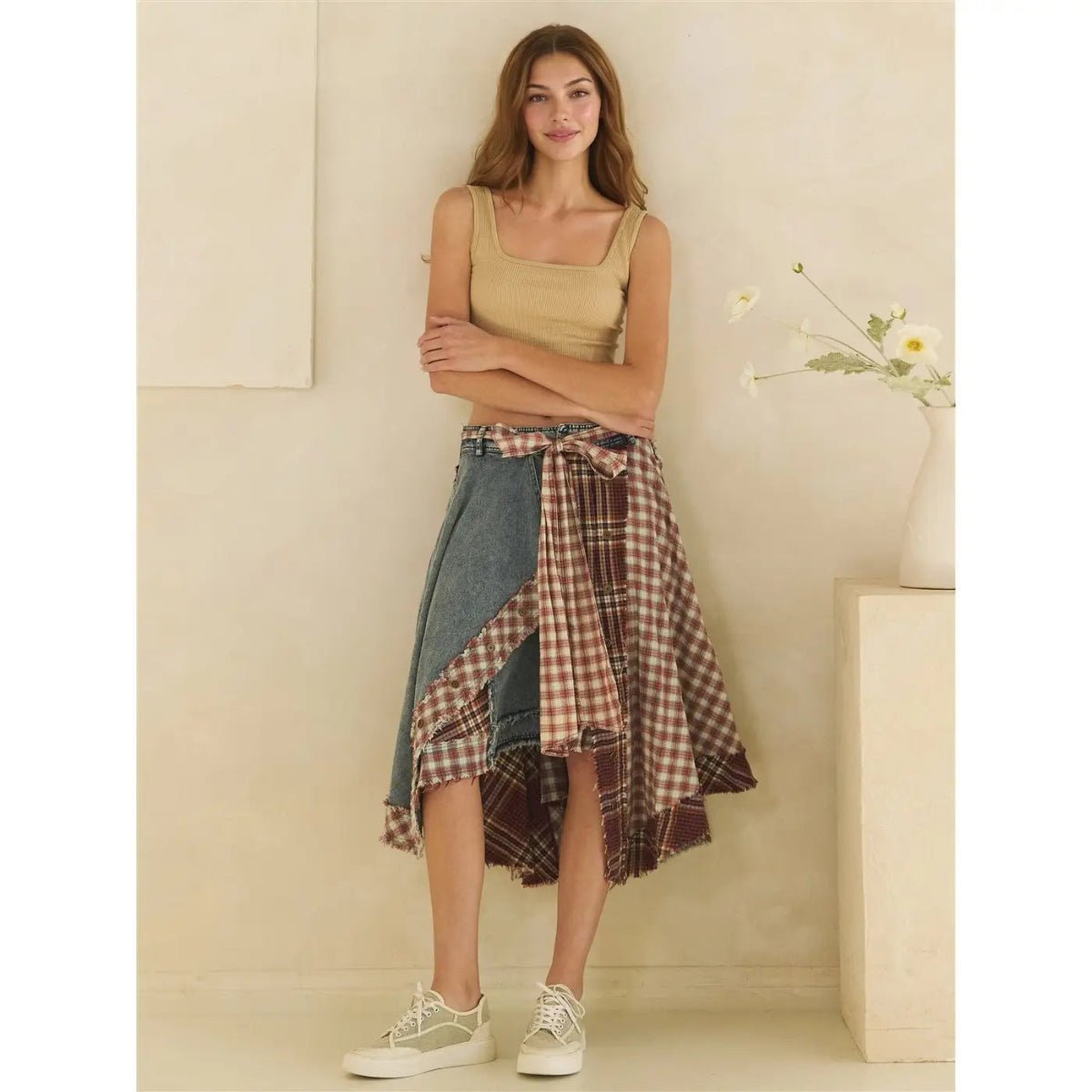 Female leaning on a wall wearing a denim skirt, beige tank top and sneakers