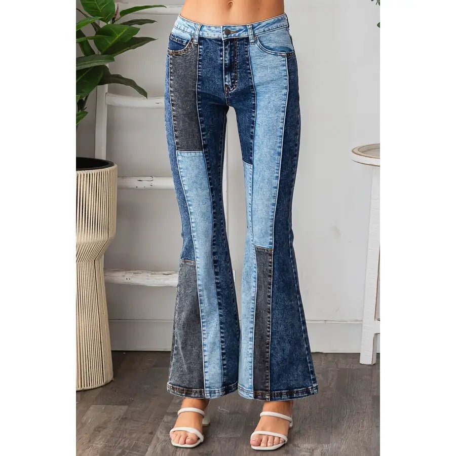 A person wearing high-waisted patchwork flare jeans made from multiple shades of blue denim, paired with white heeled sandals, standing in an indoor room setting.