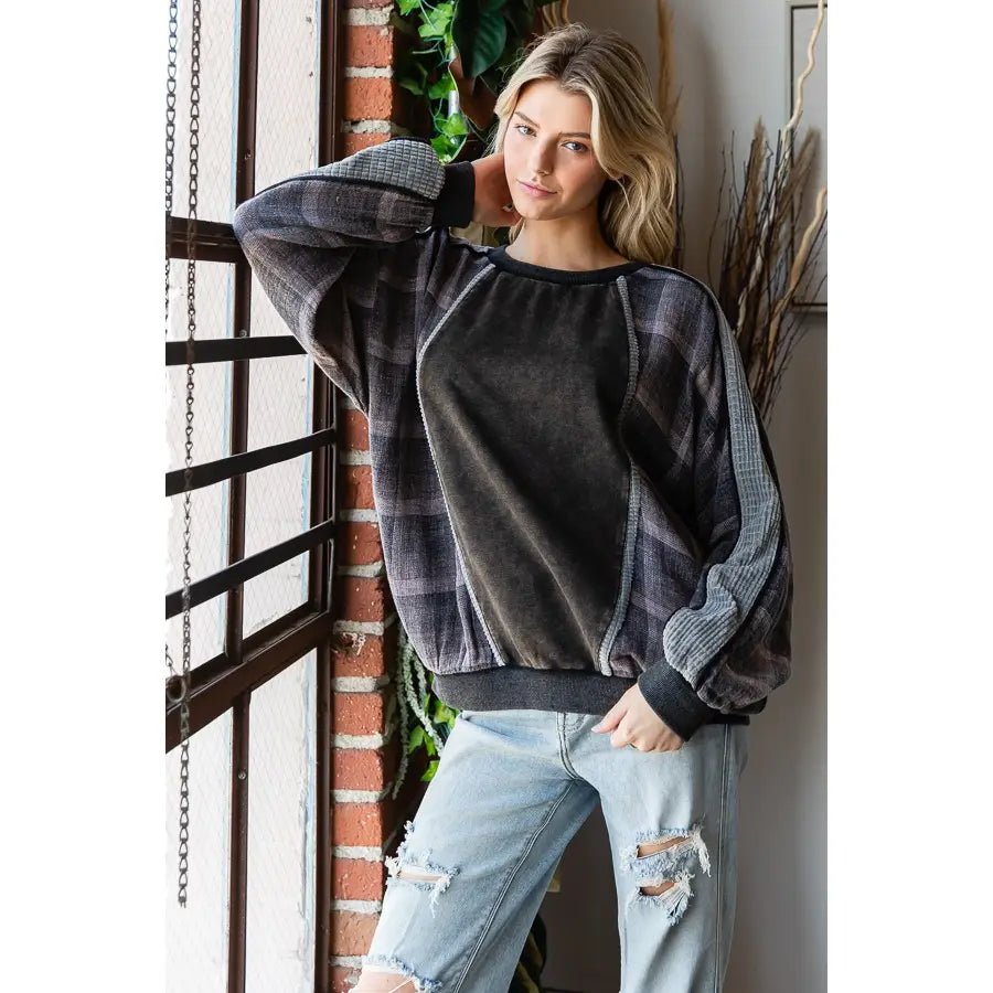 A woman models a dark gray and black plaid sweatshirt with a solid dark gray front panel and textured gray sleeves, paired with light-wash ripped jeans, standing next to a brick column.