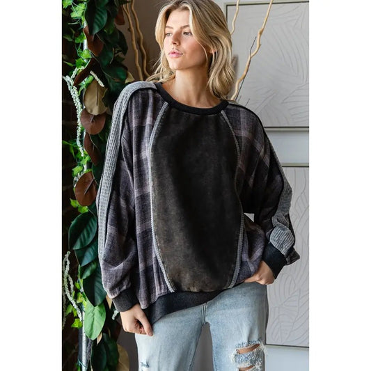 A woman wearing a dark charcoal gray and black plaid raglan sweatshirt with textured knit sleeves, paired with light-wash distressed jeans, standing in front of a paneled wall with decorative plants.