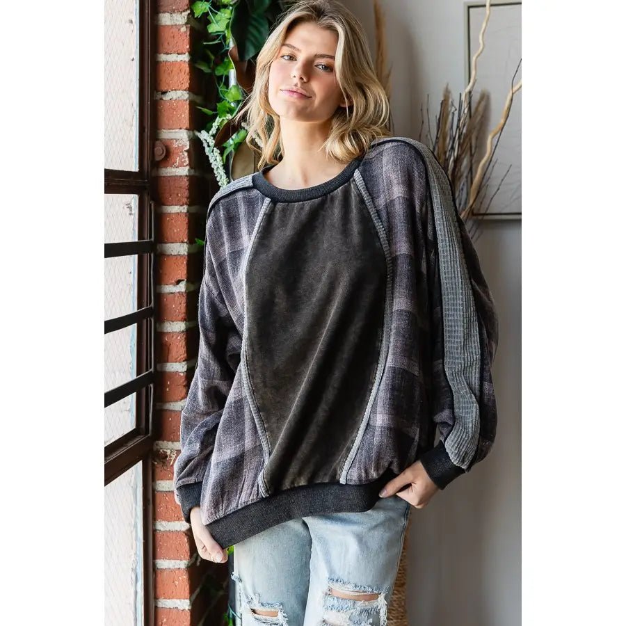 A woman wearing a dark gray and black plaid long-sleeve sweatshirt with a solid dark gray front panel, standing next to a brick column.
