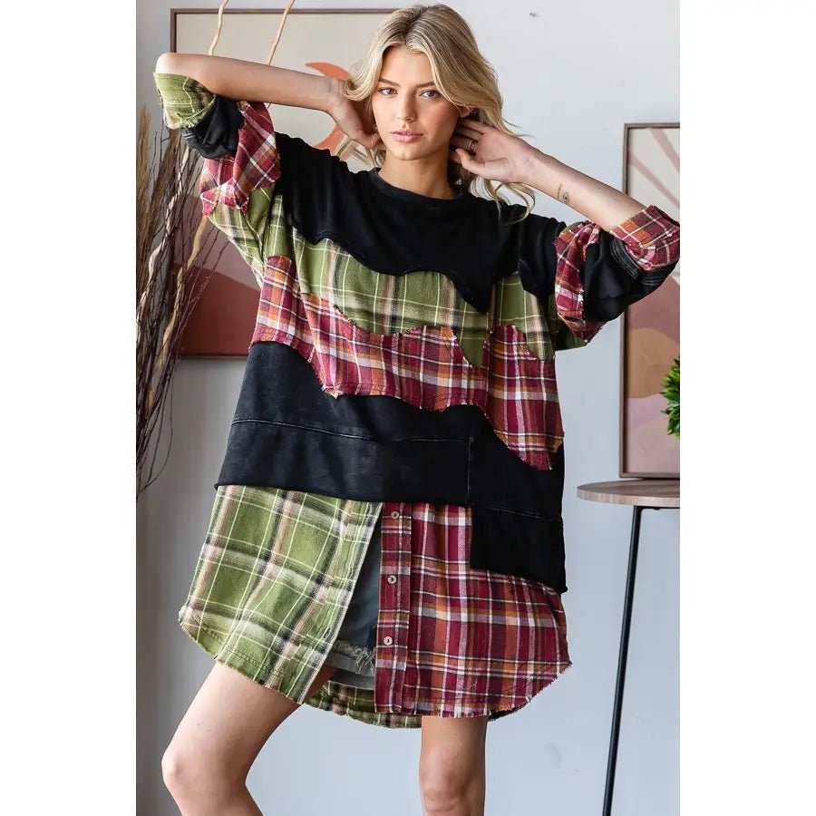 A woman modeling a patchwork shirt dress with mixed plaid and solid fabric panels in green, red, black, and cream, posed in an indoor setting.