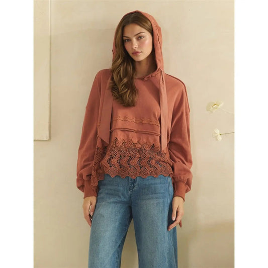 A woman modeling a rust-colored hooded long-sleeve top with a lace hem, paired with blue jeans, standing against a neutral beige wall.