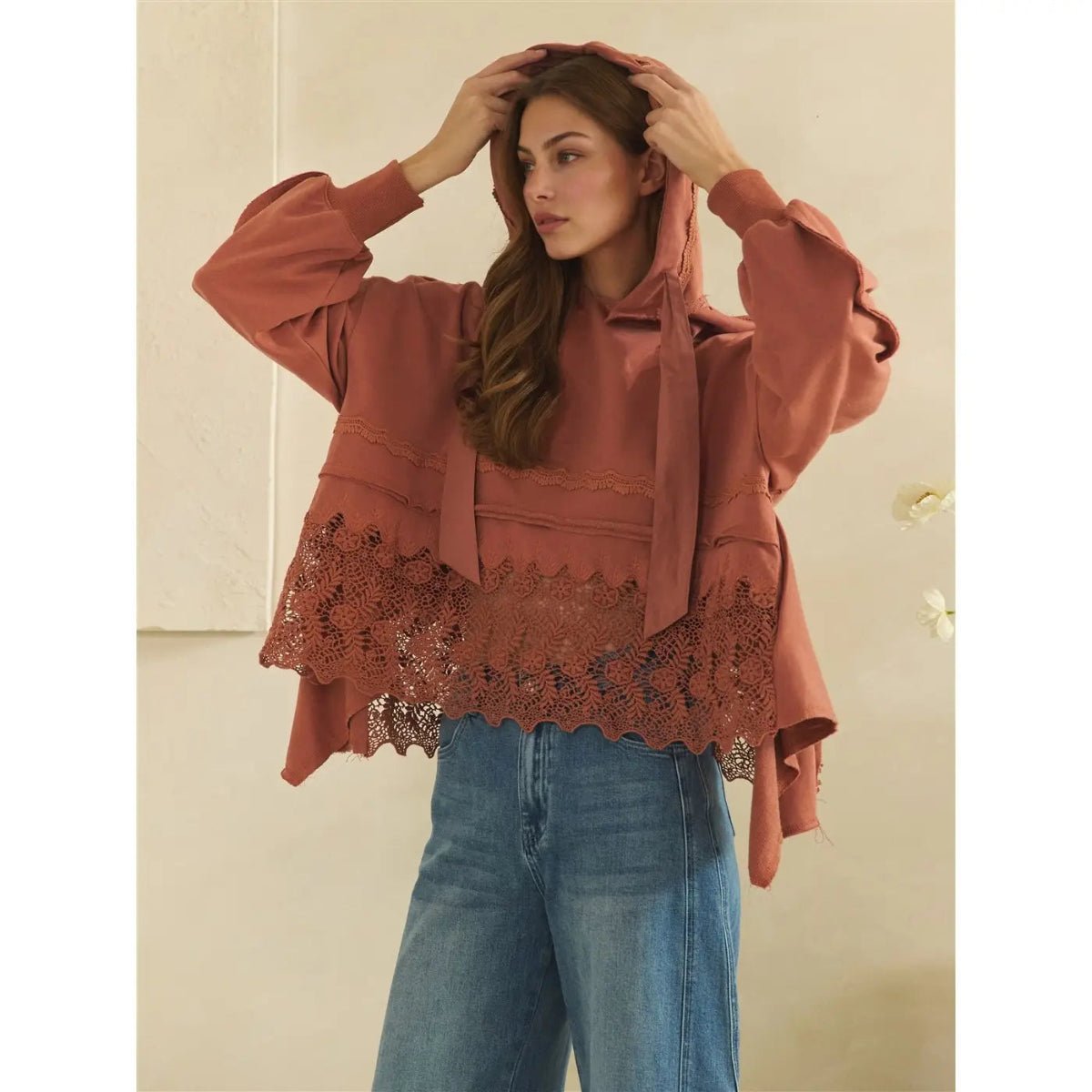 A woman wearing a terracotta rust-colored hooded sweatshirt with lace trim and voluminous sleeves, paired with blue jeans, posing against a neutral beige wall.