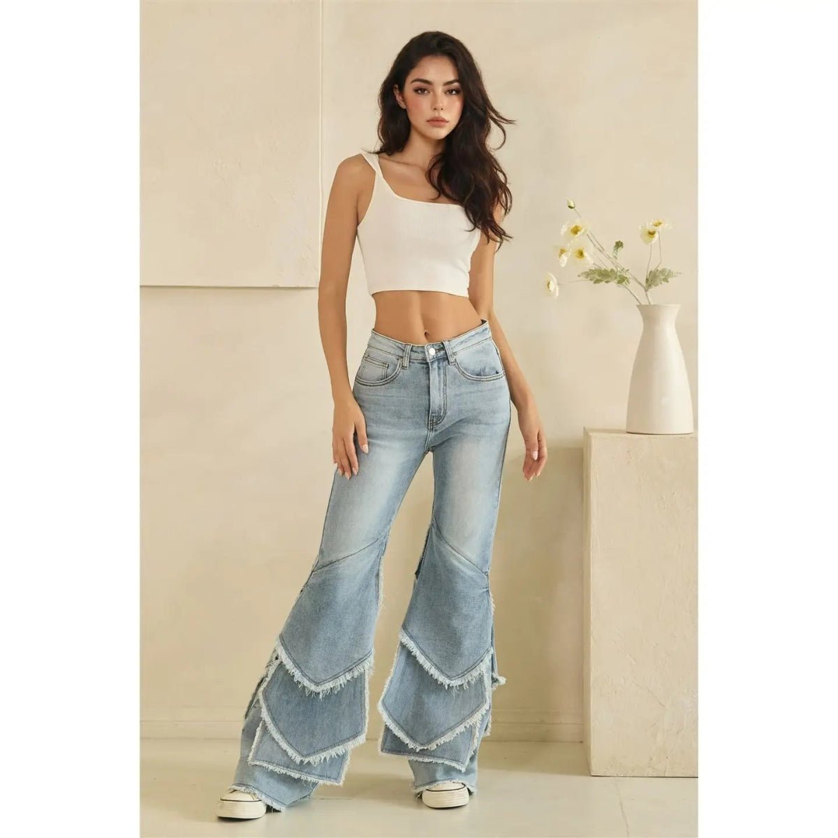 A woman stands indoors wearing light-wash wide-leg jeans with layered ruffle hems and a white cropped tank top.