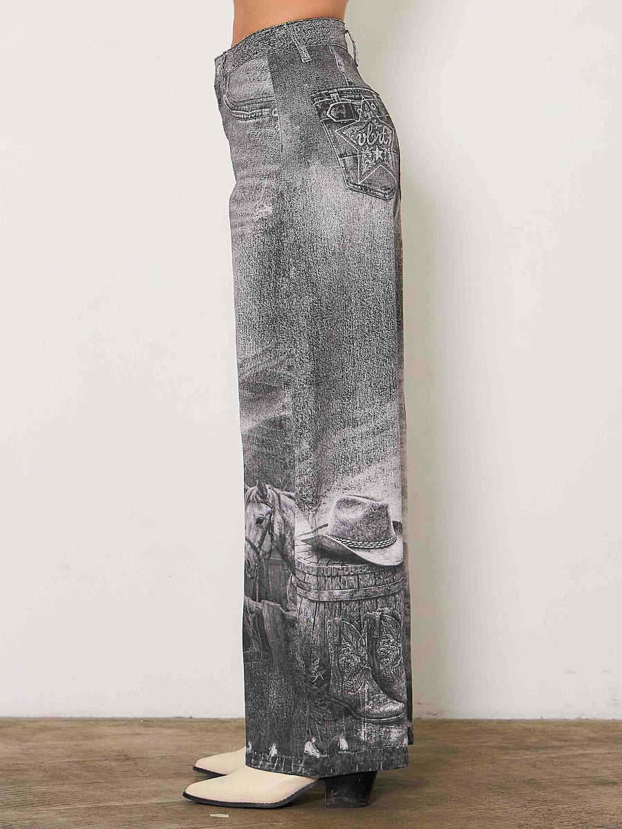 A person shown from the waist down wearing wide-leg gray denim jeans with a faded wash and western-themed graphic print near the lower legs, standing against a plain off-white wall on a wood floor.