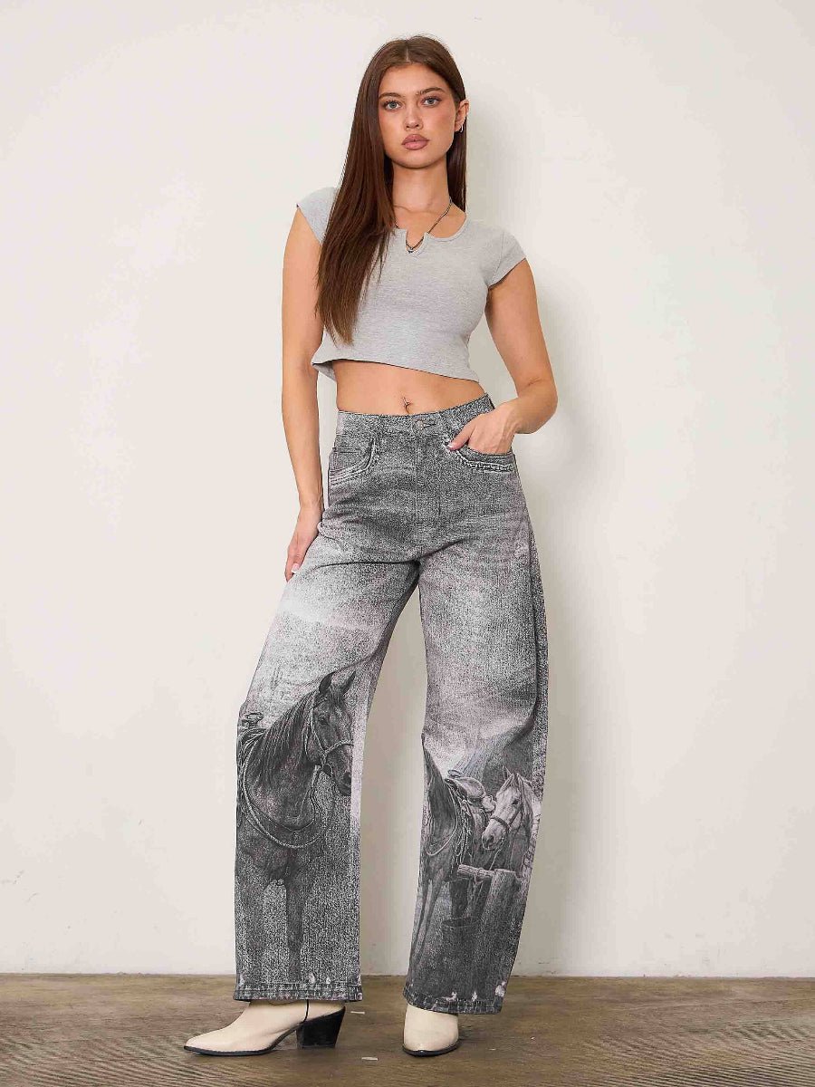 A woman modeling high-waisted, wide-leg gray jeans with a black horse graphic printed on the lower legs, paired with a light gray crop top and white pointed-toe heels, standing against a plain off-white wall.