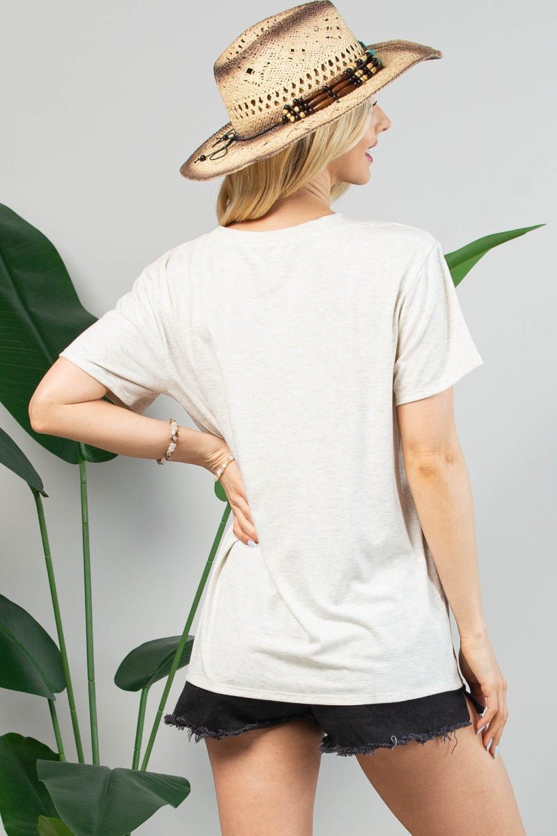 A woman shown from the back wearing a light off-white short-sleeve t-shirt, black denim shorts, and a straw hat, standing in front of a gray wall with green plant leaves.