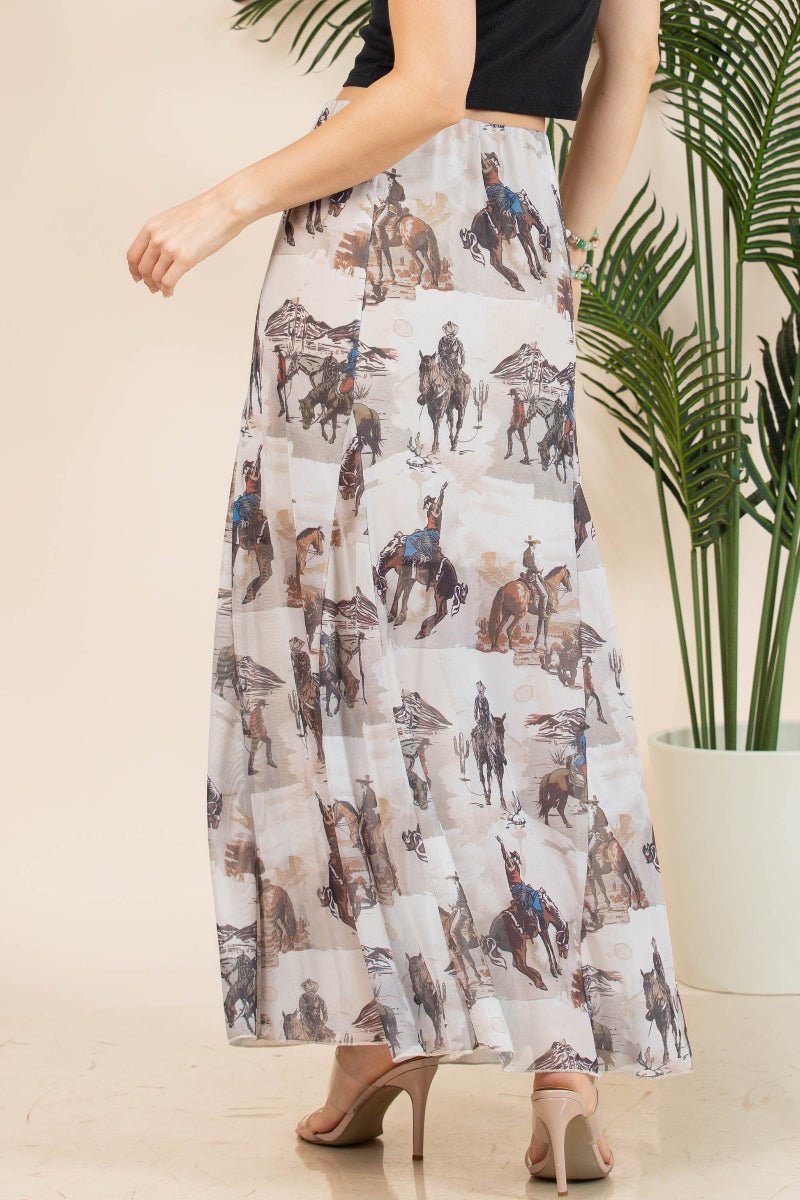 A woman wearing a long white maxi skirt with an all-over print of cowboys and horses, paired with beige high heels and a black crop top, standing in front of a beige wall with a potted plant.