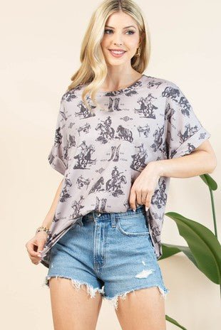 A woman modeling a short-sleeve gray t-shirt with a black cowboy and cattle print, paired with distressed denim shorts, set against a warm beige background with a green plant leaf.