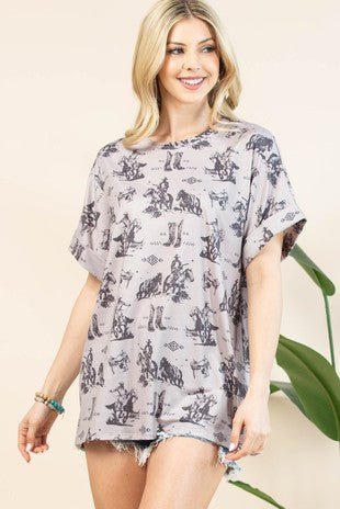 A woman modeling a light gray short-sleeve tunic top with a black cowboy and cattle print, paired with frayed denim shorts, set against a warm beige background with a green plant leaf visible on the right.